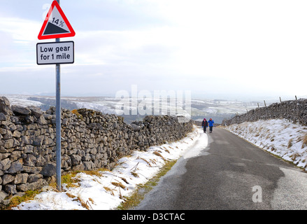 Two hikers walking up a narrow country road with dry stone wall and snow. Traffic sign warning to select low gear. Stock Photo