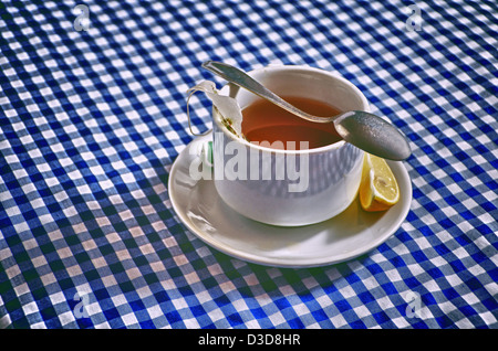 Tea. White porcelain cup of tea arranged with spoon, lemon and plate on blue checkered tablecloth. HDR toning. Stock Photo