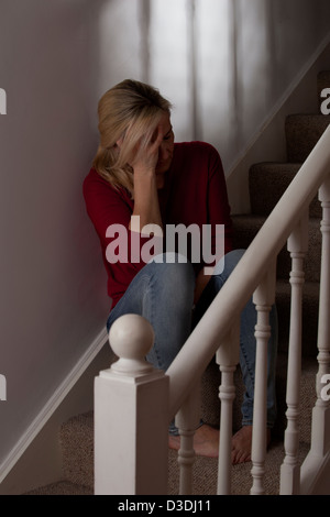 Blonde woman sitting in darkness alone on the steps, at home, hand on the side of her face depressed. Stock Photo