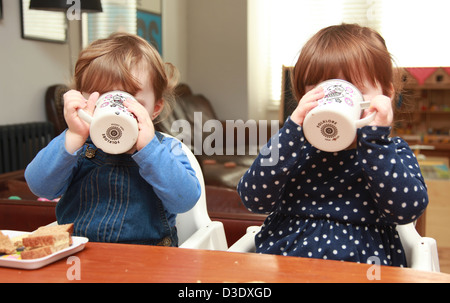 Twin girls eating sandwiches and drinking out of a cup. Stock Photo