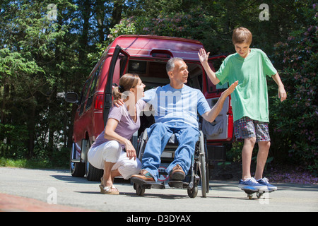 Man with spinal cord injury in wheelchair giving high-five to son on skateboard Stock Photo