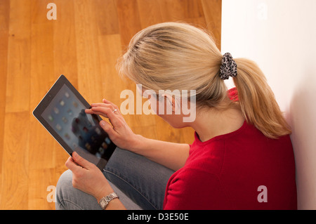 Female blonde hair in a pony-tail, sitting using a digital tablet, over the shoulder back to camera. Stock Photo