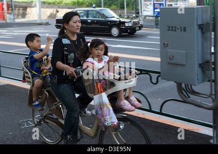 A stressed mother with her two little carefree children aboard a bicycle navigates the busy streets of downtown Tokyo. Stock Photo