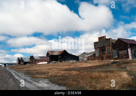 Historic wooden buildings of 'Ghost Town' under clouds in sky along wet dirt road at Bodie State Historic Park in California Stock Photo