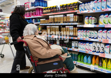 An elderly lady in a wheelchair and her carer or assistant look at hair products and shampoo in a supermarket. Stock Photo