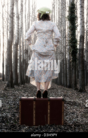 a woman in a floral vintage dress is standing on an old suitcase Stock Photo