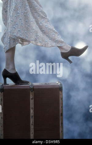 a woman in a floral vintage dress is standing on an old suitcase Stock Photo