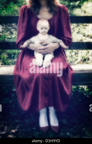 a woman in a red dress is sitting on a wooden bench with an old doll on her lap Stock Photo