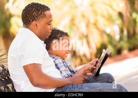 Happy African American Father and Mixed Race Son Having Fun Using Touch Pad Computer Tablet Outside. Stock Photo