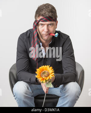 Bored businessman looking depressed at party. Sitting in armchair holding a plastic flower and wearing the tie on his forehead. Stock Photo
