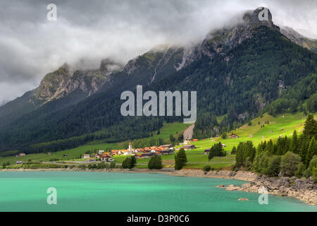 Small village on sunny valley among alpine lake with calm turquoise water and mountain peaks under cloudy sky in Switzerland. Stock Photo