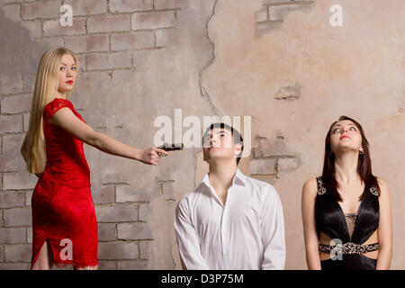 Beautiful blonde woman in a red dress threatening two young hostages who are kneeling in front of her with a handgun Stock Photo