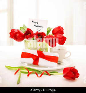 Image of fresh red tulips bouquet in beautiful vase with greeting postcard on the table, little white giftbox with ribbon, Stock Photo