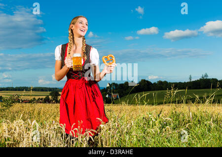 Young Bavarian woman drinking beer and keeping a pretzel in dirndl at meadow Stock Photo