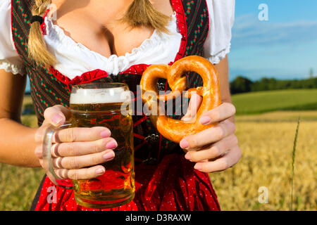 Young Bavarian woman drinking beer and keeping a pretzel in dirndl at meadow Stock Photo