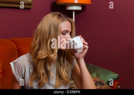 young woman sitting on a couch in an old fashioned cafe and drinks a cup of coffee Stock Photo