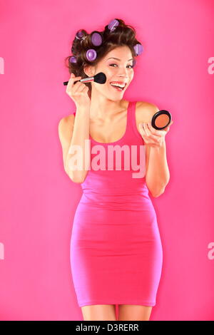 Portrait of funky trendy young mixed race woman with hair rollers in pink dress putting makeup isolated on pink background Stock Photo