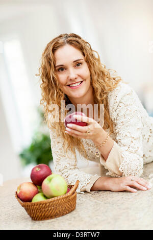 Portrait of smiling woman holding red apple leaning on countertop in kitchen at home Stock Photo