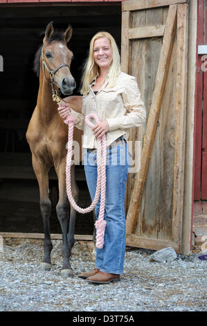 Woman leading young Arabian horse out of barn door, blond in her thirties with four month old filly. Stock Photo