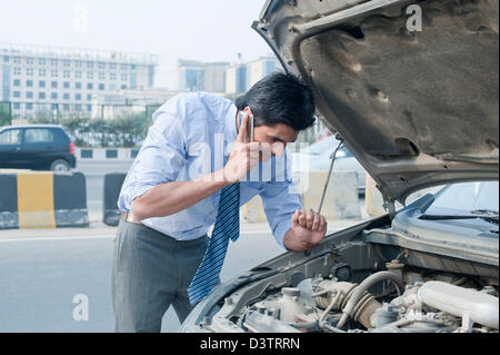 Businessman using a mobile phone near a broken down car, Gurgaon, Haryana, India Stock Photo