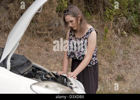 A young adult woman looks anxiously into the engine compartment of her broken down car. Stock Photo