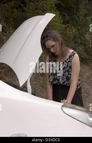 A young adult woman looks anxiously into the engine compartment of her broken down car. Stock Photo