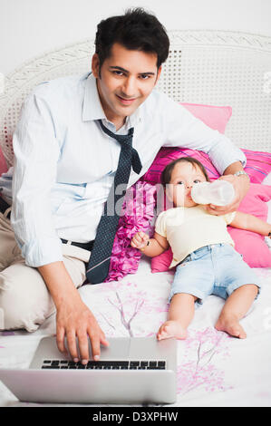 Businessman using a laptop and feeding his baby on the bed Stock Photo