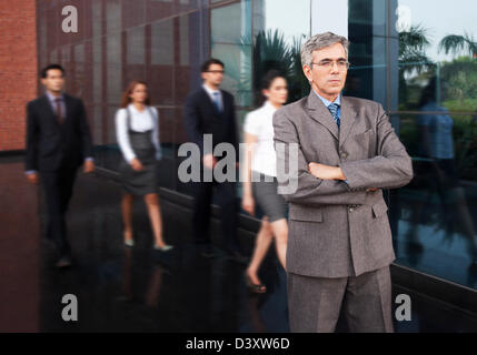 Businessman standing with his arms crossed with their colleagues walking in the background Stock Photo