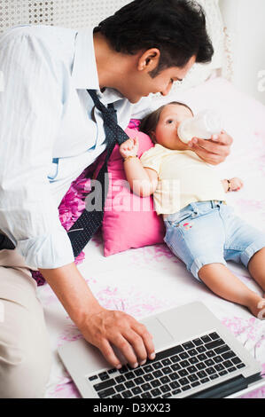 Businessman using a laptop and feeding his baby on the bed Stock Photo