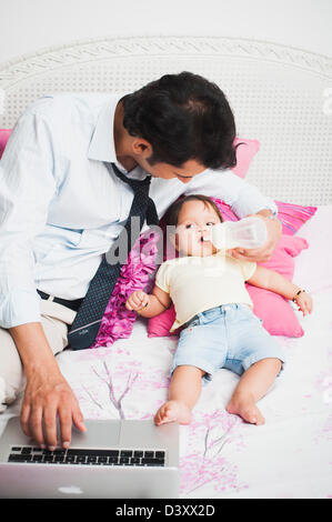 Businessman using a laptop and feeding his baby on the bed Stock Photo