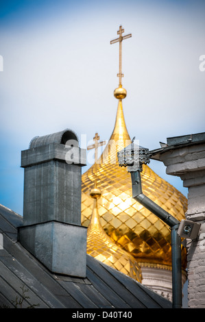 Golden domes with crosses of Russian Christian church shining behind old wooden roof with chimney. Contrast of grand and simple. Stock Photo