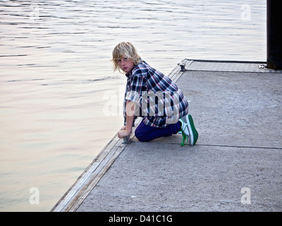 Young blond Australian boy at a jetty or pier, wearing blue check Flannelette shirt, purple skinny jeans and green skate shoes Stock Photo