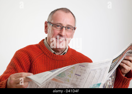 Mature man in 50s wearing sweater, reading a broadsheet newspaper, looking to camera smiling. Stock Photo