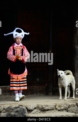 A Long Horn Miao little girl in traditional costume standing with her pet dog by her home. Stock Photo