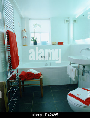Slate flooring and chrome towel rail in modern white bathroom with red towels on bamboo stool beside white tiled bath Stock Photo