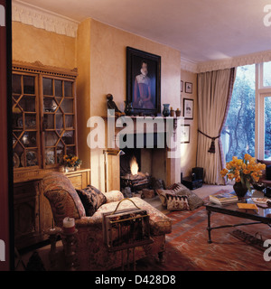 Armchair in front of glass-front dresser in living room with large painting above fireplace and white drapes on French windows Stock Photo