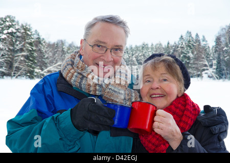 Smiling attractive senior couple outside in snowy winter landscape, holding warm cups of drinks Stock Photo