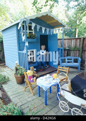 Small girl wearing dungarees standing in doorway of blue wooden playhouse on patio with children's furniture and dolls' pram Stock Photo