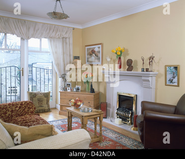 Cream sofa and brown leather chair beside fireplace in first floor apartment living room with cream curtains on French windows Stock Photo