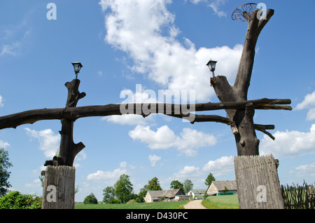 wooden park gate made of old tree trunks and rural homestead houses on blue cloudy sky. Stock Photo