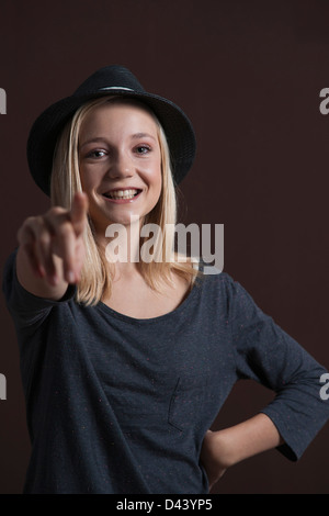 Portrait of Blond, Teenage Girl, Smiling at Camera, wearing Hat and Pointing finger, Studio Shot on Black Background