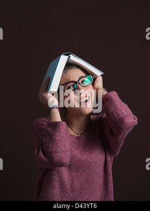 Portrait of Teenage Girl wearing Eyeglasses, holding Open Book over Head with Anxious Expression, on Black Background