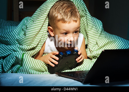Caucasian happy kid hiding under blanket playing computer games Stock Photo