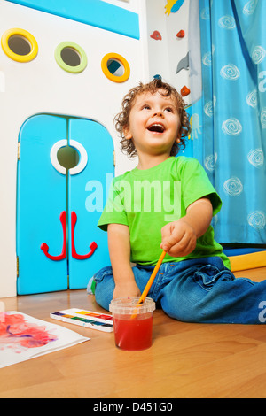 Two years old boy painting with laughing expression dipping paintbrush into water bucket Stock Photo