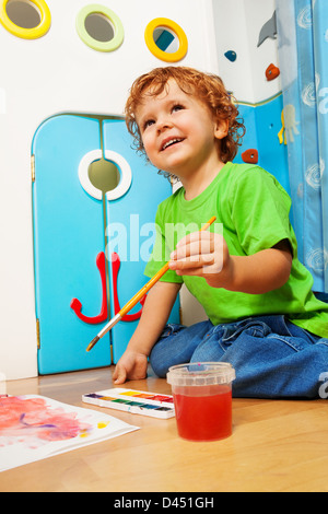 Two years old boy painting holding and dipping paintbrush into water bucket with smile on his face Stock Photo