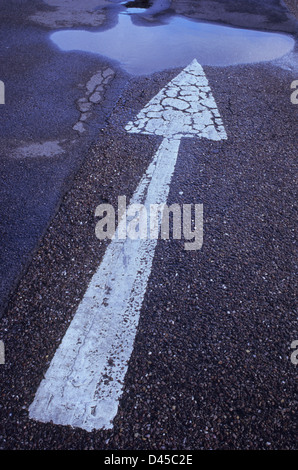 Cracked and worn arrow pointing away from viewer painted white on tarmac surface with large puddle Stock Photo