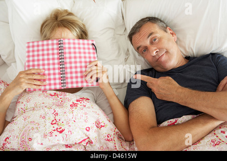 Bored Looking Senior Man Lying In Bed Next To Senior Woman Reading Book Stock Photo