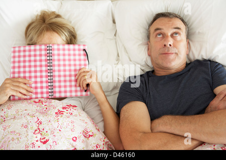 Bored Looking Senior Man Lying In Bed Next To Senior Woman Reading Book Stock Photo