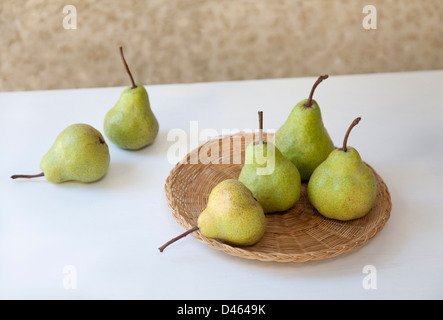 Six green pears on a woven rattan dish set on a white table top with a patterned background Stock Photo