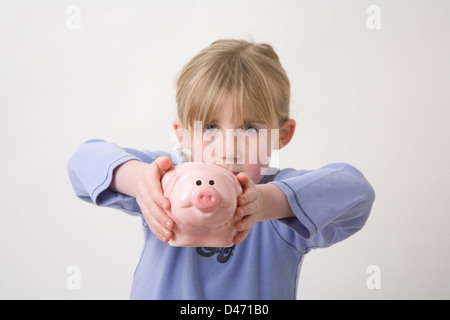 young girl with blonde hair tied back, in blue long sleeved t-shirt holding pink piggy bank towards camera Stock Photo